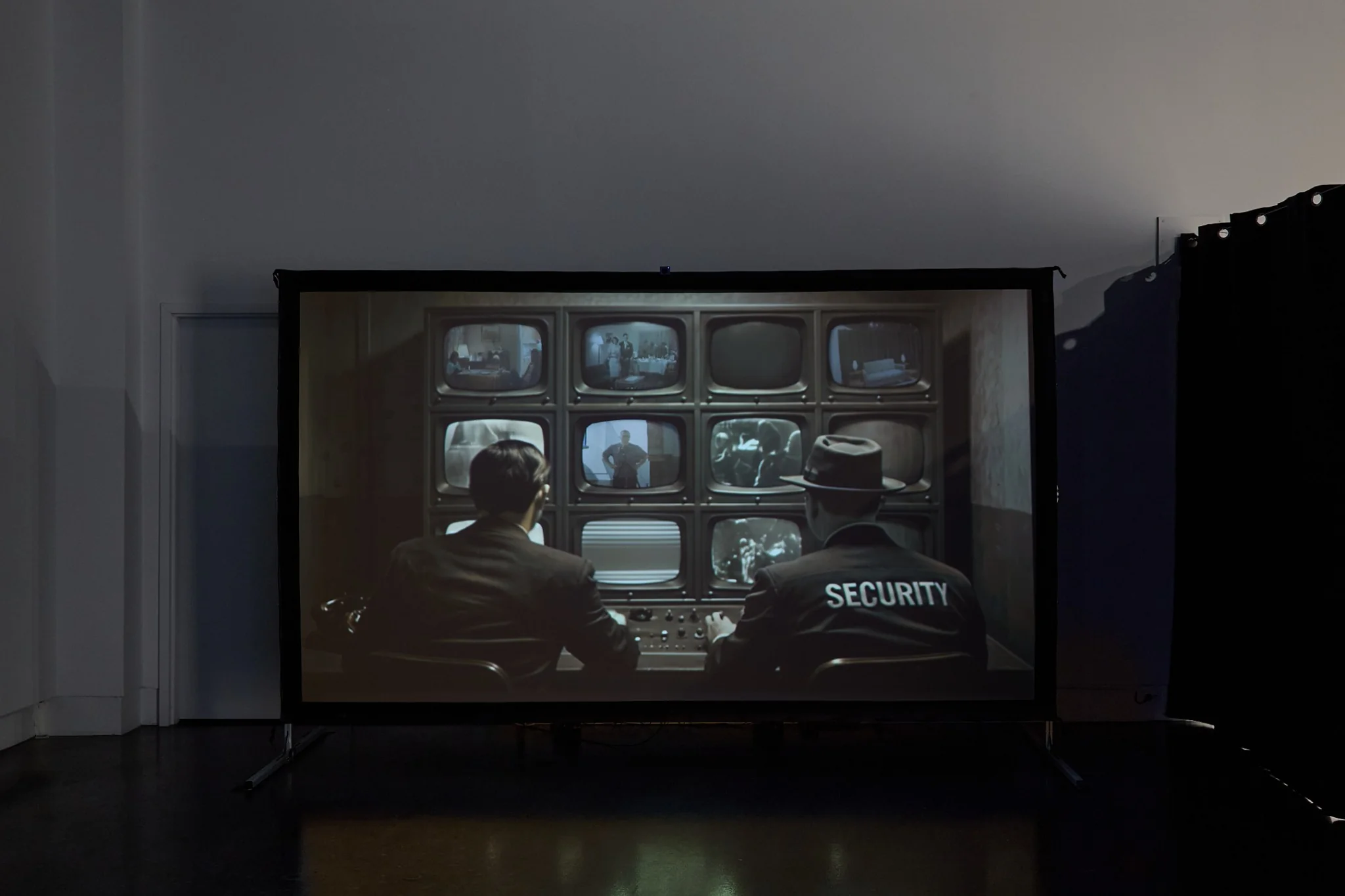 Large projected image of two security agents facing a wall of surveillance monitors
