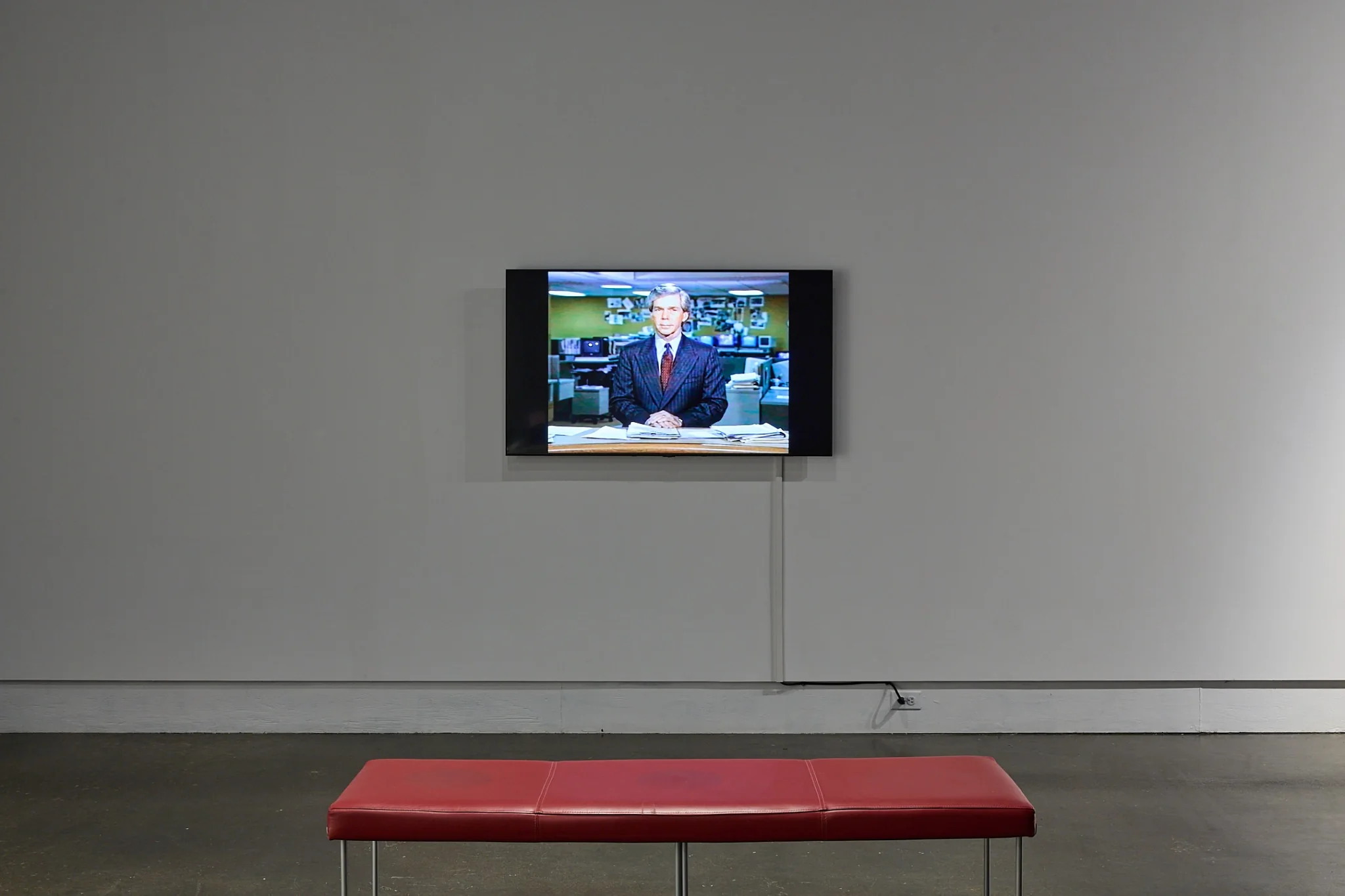Wall-mounted monitor showing an AI-generated television reporter with a red gallery bench below